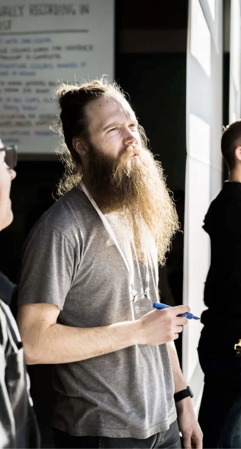 Photo of a man with a long beard and contemplative expression, wearing a grey t-shirt and a conference attendee lanyard, standing in a sunlit area.